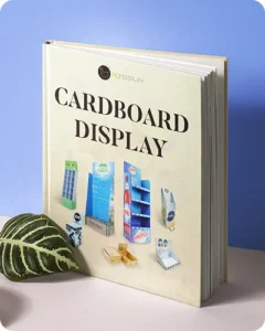 A photo of a book titled 'CARDBOARD DISPLAY,' featuring images of various cardboard display stands on the cover. The book is placed on a surface with a plant leaf nearby.