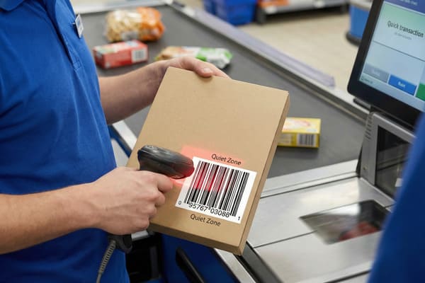 A person wearing a blue polo shirt uses a handheld barcode scanner to scan a brown cardboard box at a retail checkout counter. The scanner emits a red laser light over the barcode, which is clearly visible with the number 095767030881 and the text 