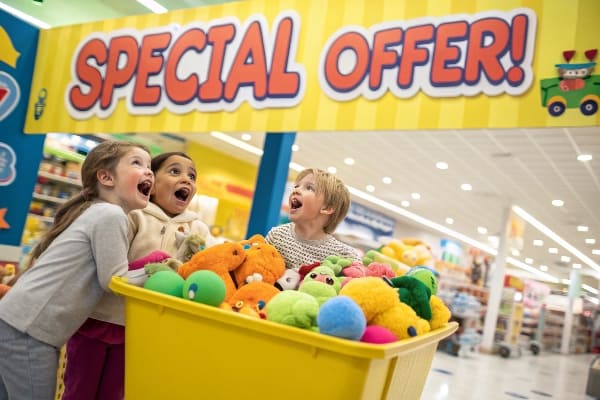 Trois enfants excités regardant un bac jaune plein de jouets en peluche sous un panneau d'offre spécial