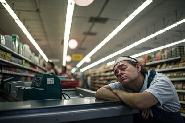 疲れた食料品店のレジ係は、蛍光灯の下でチェックアウトカウンターで頭を置いています。