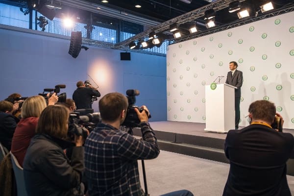 Man speaking at press event with photographers capturing the moment