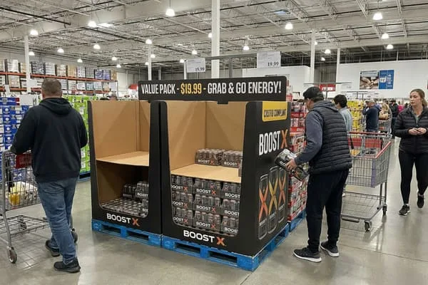 镜头拉远，展现了熙熙攘攘的Costco仓储式超市过道，画面中心是一个大型黑棕色纸板展示架，上面摆放着“BOOST X”能量饮料。展示架放置在蓝色工业托盘上，醒目地印着“超值装 - 19.99美元 - 即拿即走能量饮料！”和“符合Costco标准”的字样。货架上堆满了多罐装的Boost X能量饮料。一位身穿黑色背心和帽子的男士正伸手去拿一包Boost X。左侧，一位身穿黑色连帽衫的男士推着一辆购物车，里面装着一只小狗。右侧，一位身穿黑色羽绒服的女士走过。背景中，高高的货架上摆满了散装商品，头顶的灯光明亮。