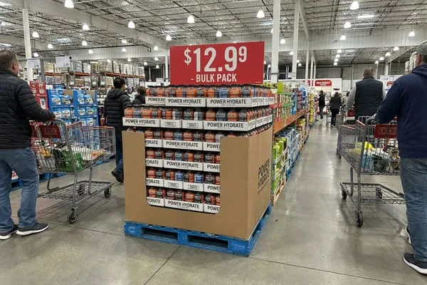 在类似好市多（Costco）的仓储式超市宽敞的过道里，一个托盘上醒目地摆放着一大堆Power Hydrate运动饮料，橙色、蓝色和黑色的瓶子格外引人注目。货架上方一块鲜红色的标牌醒目地写着“大包装”，售价12.99美元。几位顾客推着满满的购物车在熙熙攘攘的过道里穿梭，背景是高高的天花板、工业照明和摆满其他散装商品的货架。