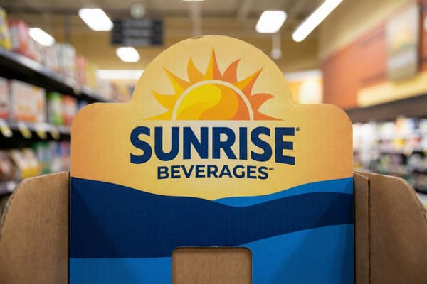 A vibrant cardboard point-of-sale display for 'Sunrise Beverages' in a supermarket aisle. The display features a prominent yellow and orange sun logo at the top, with the brand name 'SUNRISE BEVERAGES' in dark blue text. Below the text, there are stylized blue waves. The background is blurred, showing rows of products on shelves and overhead lighting, indicating a retail environment.