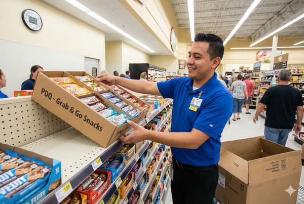 青いポロシャツを着て名札を付けた笑顔の男性食料品店従業員が、段ボールを慎重に置いている。 