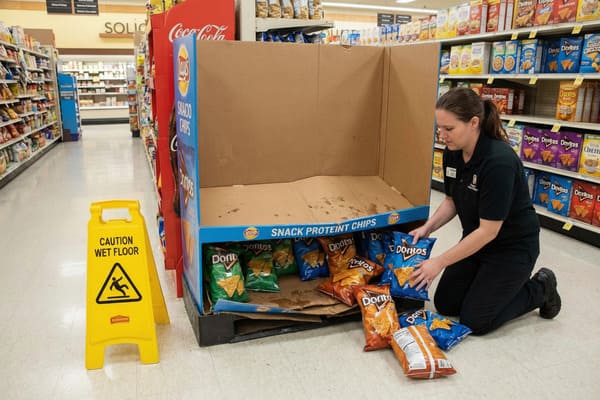 Une employée de supermarché en uniforme noir s&#39;agenouille pour ranger des paquets de chips Doritos dans un présentoir de snacks Lay&#39;s, dans une allée. 