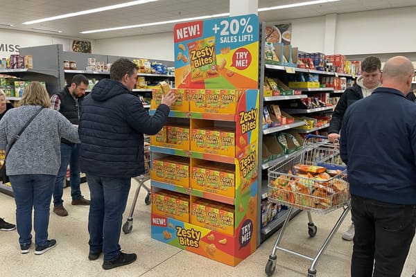 A man in a blue jacket examines a package of 'Zesty Bites' snacks from a large, colorful promotional display in a busy supermarket aisle. The display advertises 'NEW!' and '+20% SALES LIFT!' for the snack. Another shopper's cart is filled with multiple 'Zesty Bites' packages, indicating popularity. Other shoppers are visible in the background, browsing shelves.