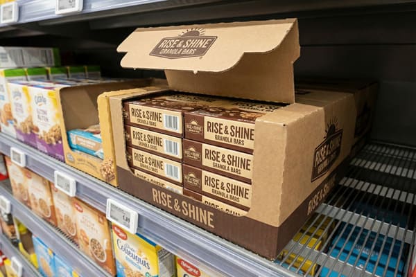 An open brown cardboard display box of Rise & Shine Granola Bars, featuring brown and beige packaging, is prominently displayed on a metal wire shelf in a grocery store aisle, with individual granola bar boxes visible inside. Other breakfast and snack items are blurred in the background.