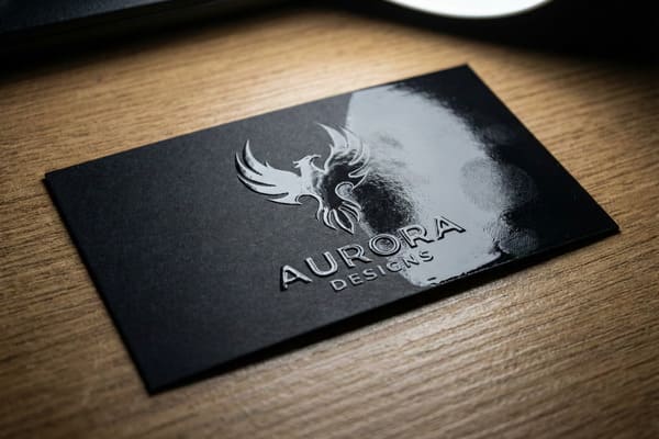 صورة مقرّبة لبطاقة عمل سوداء فاخرة تحمل شعار طائر الفينيق البارز بتقنية الأشعة فوق البنفسجية اللامعة، بالإضافة إلى عبارة "AURORA DESIGNS". تتميز البطاقة بسطح غير لامع على ورق مقوى أسود، مما يخلق تباينًا مع العناصر اللامعة البارزة. تستقر البطاقة على مكتب خشبي دافئ اللون ذي ملمس مميز، حيث يعكس الضوء الساطع التفاصيل اللامعة ويُظهر آثار بصمات أصابع خفيفة.