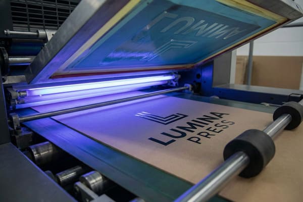 最新の工業用スクリーン印刷機のクローズアップ。茶色のクラフト紙に印刷された「LUMINA PRESS」のロゴを硬化させています。青色のUVランプが濃い黒のインクを照らし、速乾性と耐久性を確保しています。自動コンベアシステムの上に設置された、隆起したスクリーン印刷フレームからは、ロゴのステンシルが反転して見え、効率的なカスタムパッケージやブランディング資材の製造の様子が見て取れます。