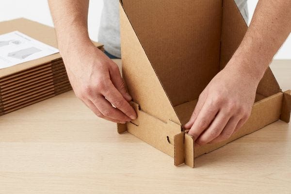 Hands assemble a brown cardboard display, interlocking structural tabs at the base on a wooden table, with flat-packed units nearby.