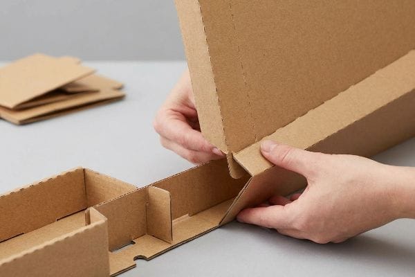 双手正在组装一个棕色瓦楞纸板展示架，折叠预先压痕的面板，并将互锁卡扣扣合在一起，形成一个牢固的底座。.