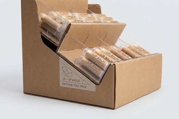 Expositor de cartón kraft marrón con alimentación por gravedad, lleno de tubos de bálsamo labial, que muestra una etiqueta con un ángulo de 12 a 18 grados y la indicación "Prueba de fricción: superada".