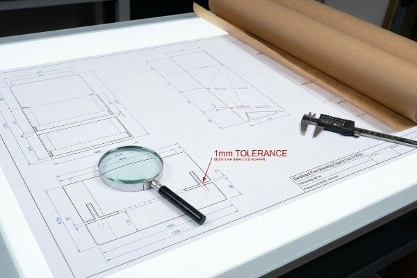 A magnifying glass rests on a technical drawing showing 1mm Tolerance for Cardboard Free Standing Displays (Fsdu), alongside a digital caliper.