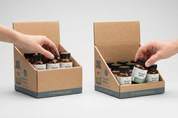 Brown corrugated cardboard display box features a 3-inch front retaining lip, filled with amber vitamin bottles, as a hand reaches inside.
