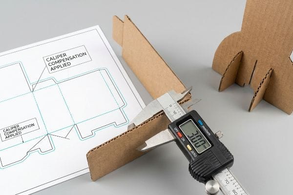 Digital caliper measuring 3.00mm corrugated board thickness, with a dieline design blueprint showing Caliper Compensation Applied.