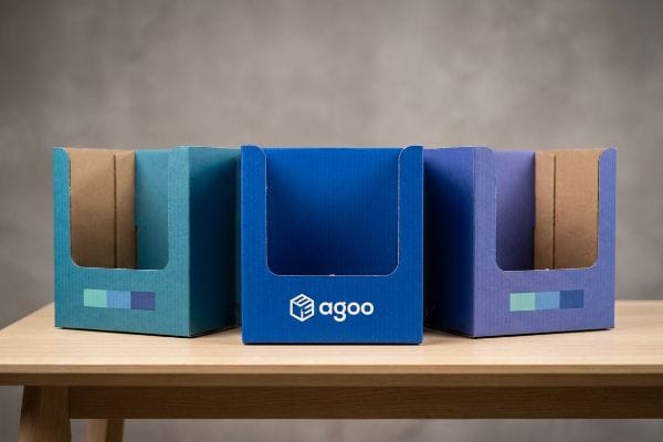 Three corrugated cardboard display boxes, in teal, royal blue with an Agoo logo, and lavender, sit on a wooden table.