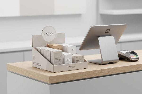 White cardboard Analoum POS display with 'Healing Soothing' and 'Simple Calm' products, next to a silver Square POS system on a wood counter.