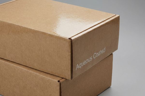 Two stacked brown corrugated boxes, the top box featuring a glossy aqueous coating and 'Aqueous Coated' text on its side.