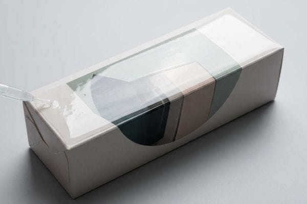 A pipette applies clear aqueous coating to a rectangular paperboard box, showcasing the water-based protective finish on its modern geometric design.