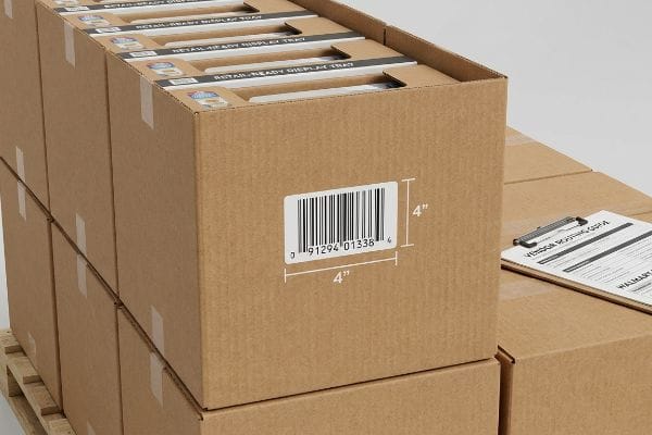 Brown corrugated master carton displaying a 4-inch flat clearance zone for a UCC-128 barcode (0 91294 01338 4), with 'Retail-Ready Display Tray' visible and a Vendor Routing Guide clipboard.