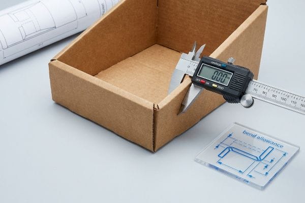 A digital caliper measures the thickness of a corrugated cardboard box, alongside a 'bend allowance' blueprint.