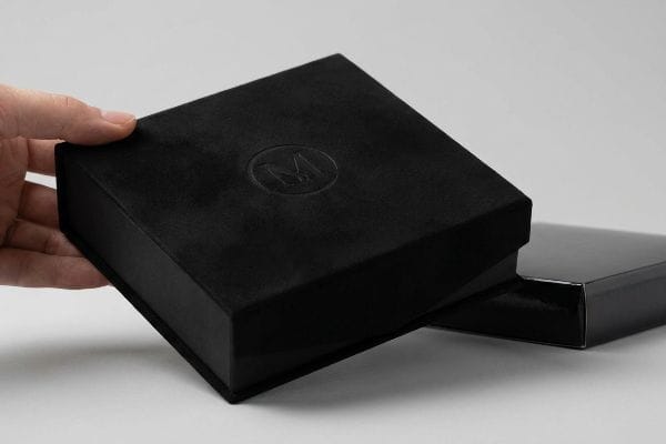 Hand lifting black velvet soft touch box with debossed M logo, showcasing premium packaging.
