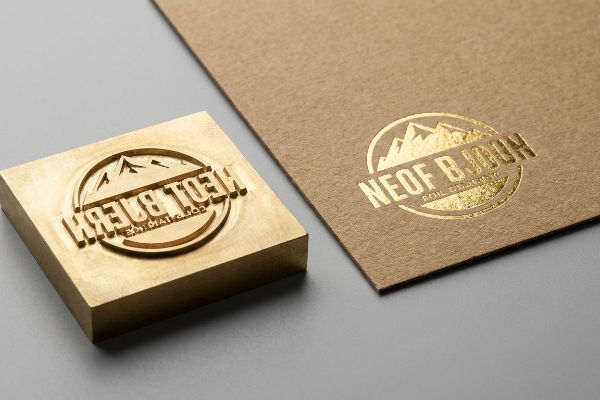 Brass foil stamping die next to brown kraft paper with a shiny gold foil Neof B. Bijouw logo, showcasing the metallic film application.