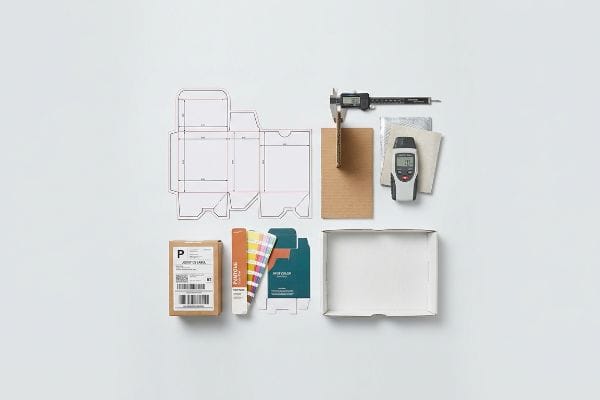Packaging quality control tools: die-cut template, digital caliper measuring corrugated board, moisture meter, Pantone swatches, and shipping box.