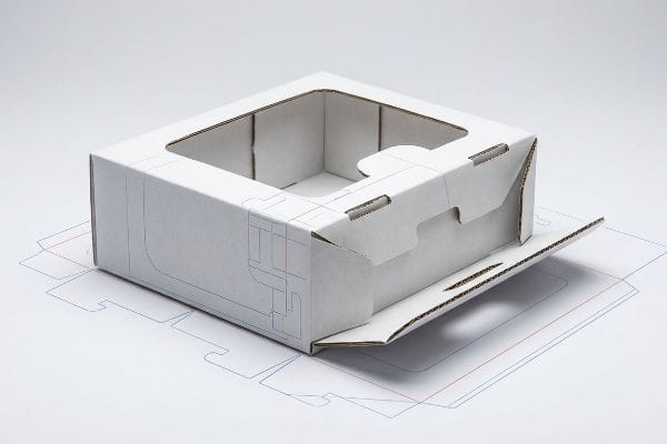 Partially assembled white cardboard packaging dieline with a square window, resting on a paper template, showcasing board caliper allowances and fold lines.
