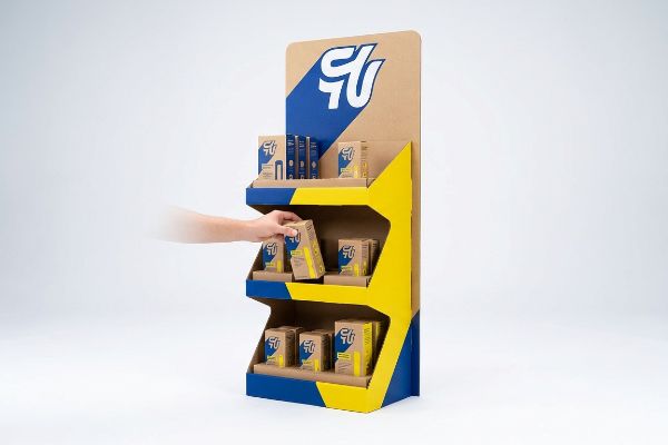 Expositor POP de cartón kraft con detalles en azul y amarillo, con el logotipo de GU, mientras una persona selecciona una caja de producto del estante central.