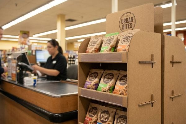 An der Kasse eines Supermarktes steht ein Pappaufsteller mit verschiedenen Snacktüten der Marke Urban Bites; im Hintergrund ist eine Kassiererin zu sehen.