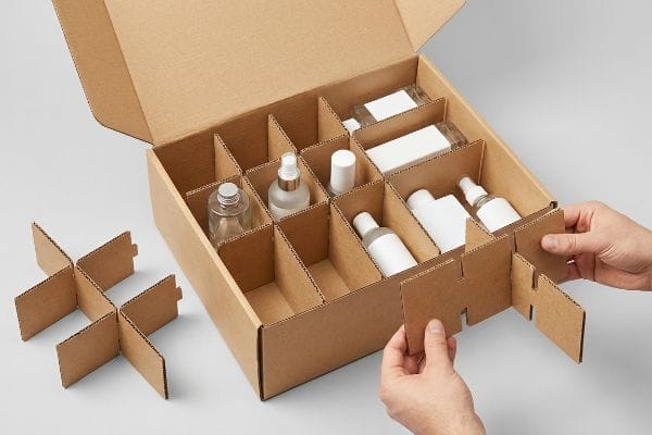 Corrugated cardboard modular dividers being adjusted by hands inside a brown shipping box, organizing various cosmetic bottles for flexible packaging.
