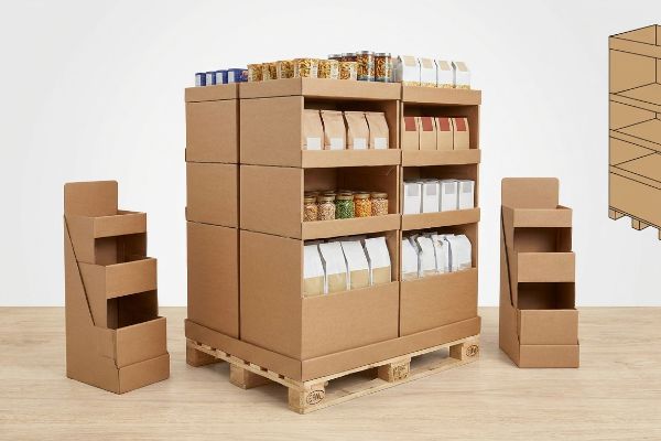 A large cardboard pallet merchandiser, stocked with various packaged food items, sits on a wooden pallet, flanked by two smaller empty retail displays.