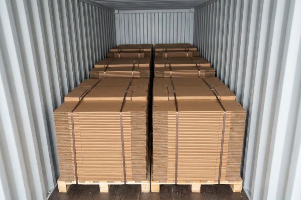 Inside a shipping container, numerous stacks of flat-packed cardboard display stands are neatly arranged on wooden pallets, optimizing freight density.