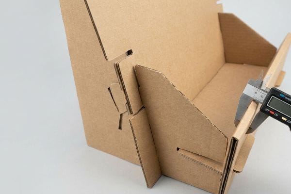 Digital caliper measuring the precise thickness of a brown corrugated cardboard display stand's interlocking tab for glueless assembly.