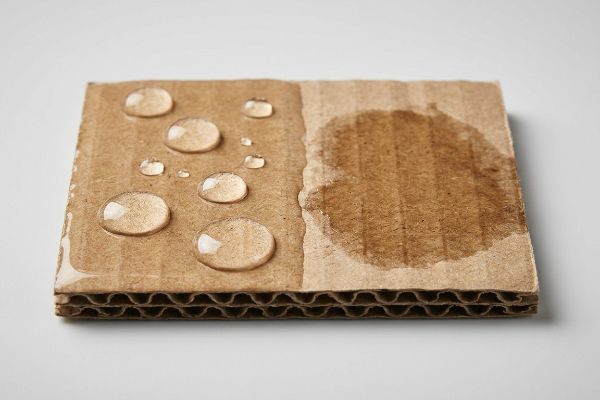 Carton ondulé avec des gouttelettes d'eau perlant sur un revêtement polymère aqueux hydrophobe, contrastant avec une tache humide et absorbée.