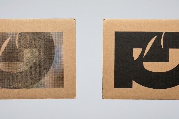 Duas amostras de papelão ondulado marrom claro, uma mostrando um gráfico preto com pontos de meio-tom CMYK visíveis em testliner poroso, a outra com uma impressão sólida em cor preta.