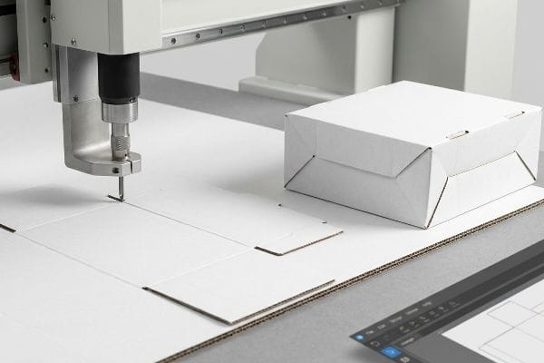 White corrugated cardboard being scored by a CNC digital cutting table, producing a white sample box, with design software for rapid prototyping.
