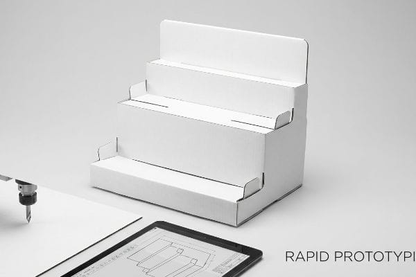 White three-tiered cardboard display prototype, precision-cut by CNC, with tablet showing CAD geometry for Rapid Prototyping.