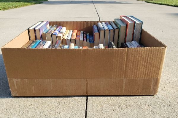 Brown corrugated cardboard box filled with numerous books displayed spine-up on concrete, ideal for a garage sale to protect from moisture.