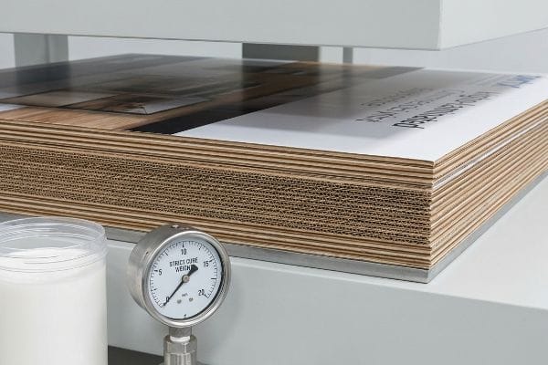 Corrugated litho-laminated sheets under pressure, beside a Strict Cure Weight gauge and PVA glue, for curing.