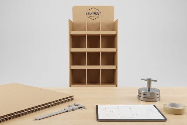 Kraft corrugated cardboard display stand with Brinikout Design Lab logo, alongside a caliper, dieline on a tablet, and raw corrugated sheets.