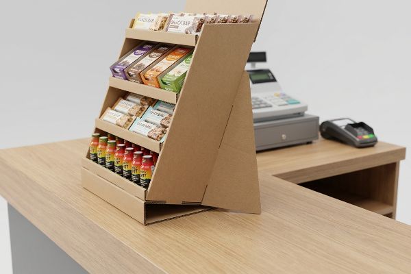 Brown corrugated cardboard countertop display featuring four tiers of Snack Bars, Energy Bars, and Hot Shot drinks on a wooden checkout counter, with a stable easel back.