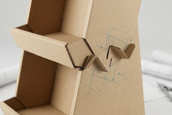 Brown corrugated cardboard display with origami-style paper locks. Blue blueprint lines detail structural bend allowance measurements.