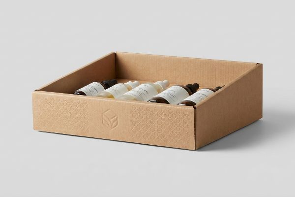 PDQ-Displaytablett aus brauner Wellpappe für sechs Glasflaschen mit Hautpflegeprodukten, verziert mit einem geprägten Muster und Logo.