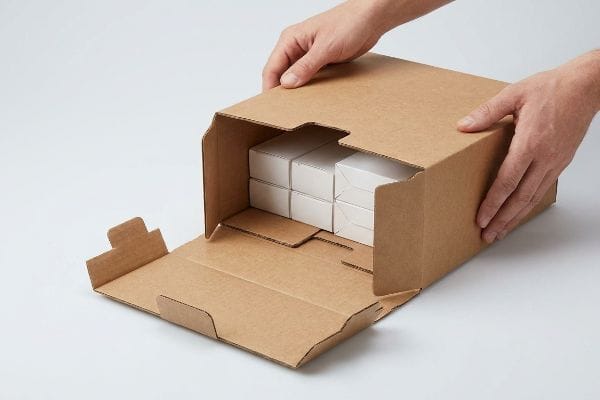 Brown corrugated cardboard PDQ packaging being opened, revealing six white modular product boxes pre-loaded inside for retail-ready display.