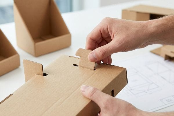 Expositor de cartón corrugado marrón que se está montando a mano, mostrando una pestaña de encaje que se inserta en una ranura.