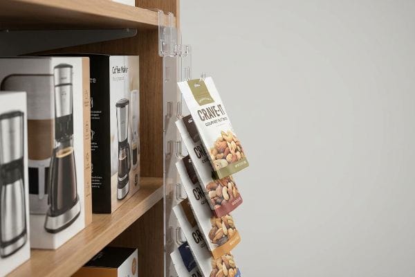 Uma faixa de plástico transparente está pendurada em uma prateleira de madeira, exibindo os pacotes de Crave-It Gourmet Nut Mix ao lado das caixas de cafeteira.