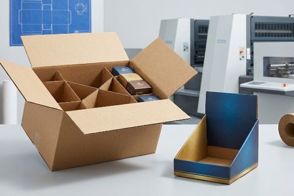 Corrugated custom box with internal dividers and small product boxes, beside a blue and gold litho-laminated retail display on a white table.